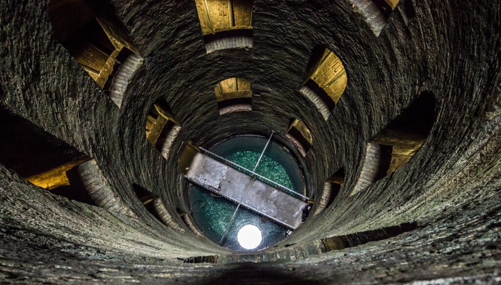 Sguardo dall’alto verso il fondo del pozzo. Sulle mura si vedono alcune finestre mentre sul fondo il ponte in ferro sospeso sull’acqua.