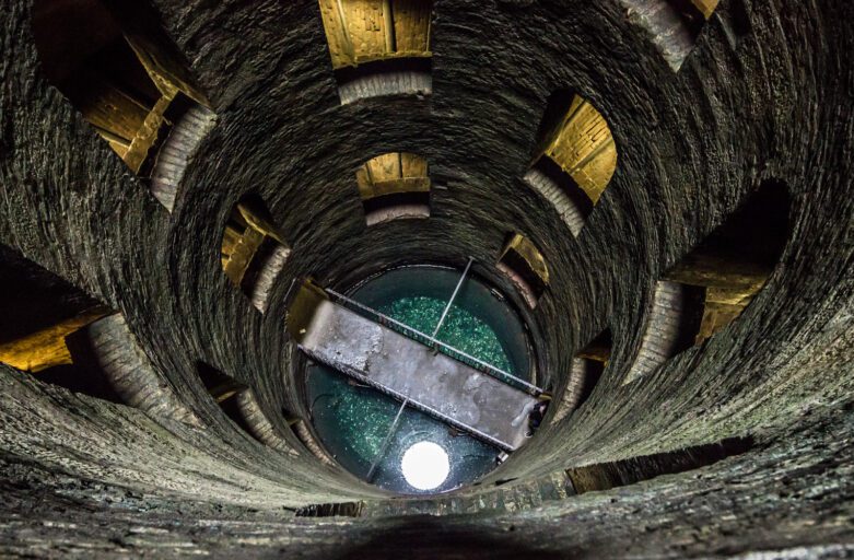 Sguardo dall’alto verso il fondo del pozzo. Sulle mura si vedono alcune finestre mentre sul fondo il ponte in ferro sospeso sull’acqua.