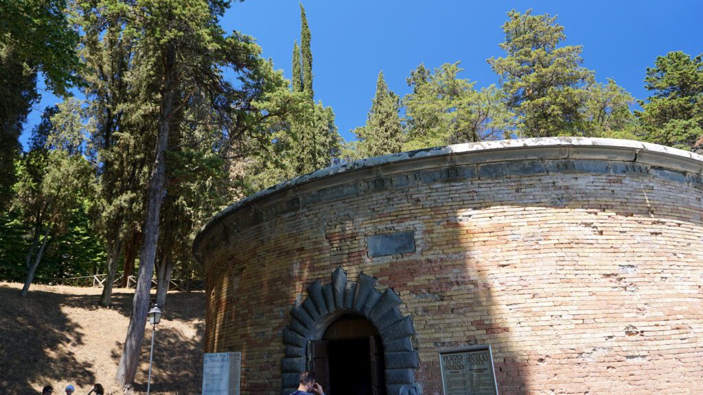 Vista dell’ingresso del Pozzo di San Patrizio. Una costruzione circolare in mattoni emerge da terra, e vi si trova il portone di accesso.