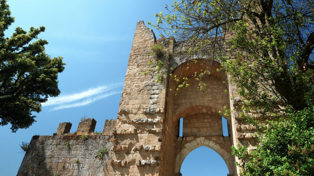 Vista dal basso della sommità del portale di ingresso al giardino municipale nella Rocca Albornoz. La muratura in mattoni, con i suoi merli, si staglia sul cielo azzurro.