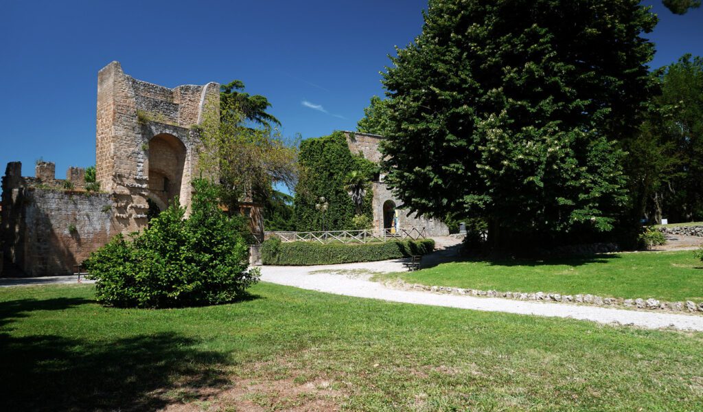 Vista del giardino municipale interno alla Rocca Albornoz, percorso da una stradina, e del portale di ingresso in mattoni.