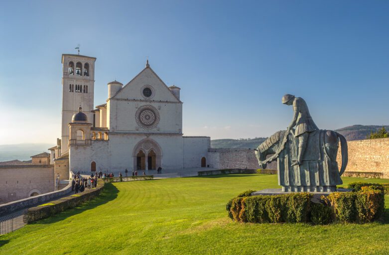 Vista della Basilica superiore di Assisi. In primo piano la statua bronzea di san francesco a cavallo sull'aiuola ben curata. Sullo sfondo la facciata della Basilica Superiore e dietro la torre campanaria.