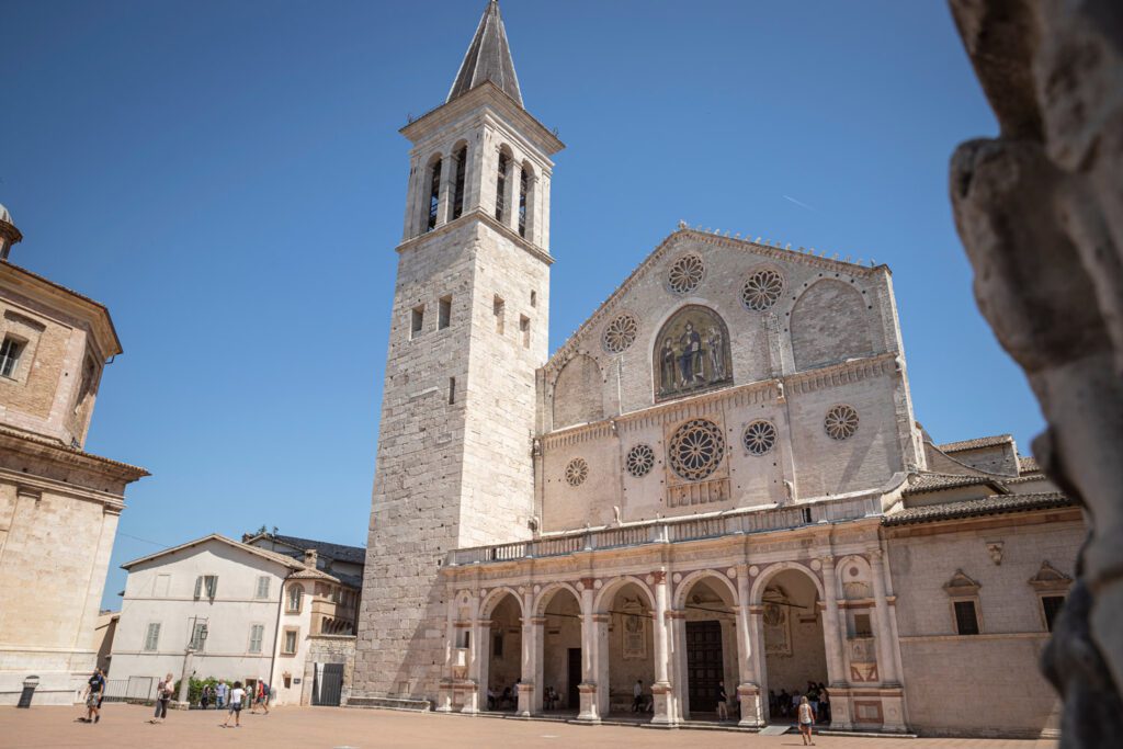 Vista scorciata dal basso a destra della facciata della cattedrale di Spoleto. La facciata e la torre campanaria si stagliano sul cielo azzurro.