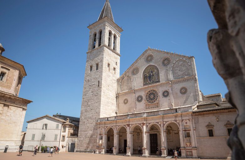 Vista scorciata dal basso a destra della facciata della cattedrale di Spoleto. La facciata e la torre campanaria si stagliano sul cielo azzurro.