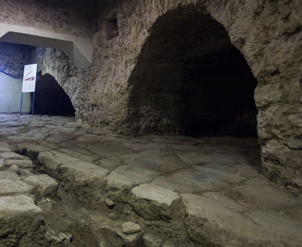 Vista della strada romana. Il decumano, realizzato con grandi pietre, è attraversato dai solchi lasciati dai carri nel tempo. Sul lato della strada romana si ergono arconi medievali utili a reggere le strutture sovrastanti.