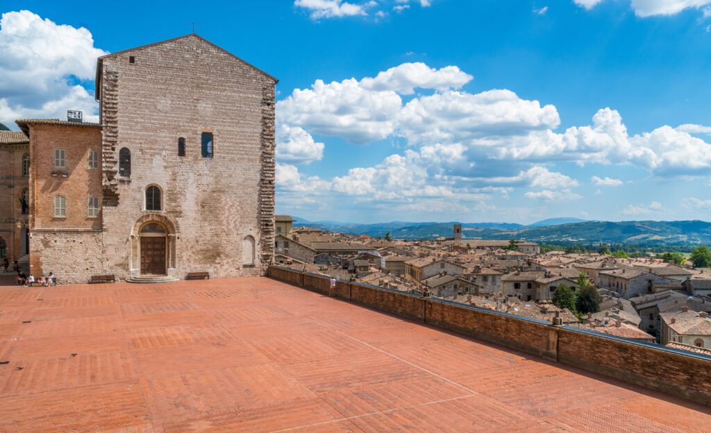 Vista di Piazza Grande dando le spalle a Palazzo dei Consoli. La piazza rossa si affaccia sui quartieri medievali sottostanti e sulle campagne umbre.