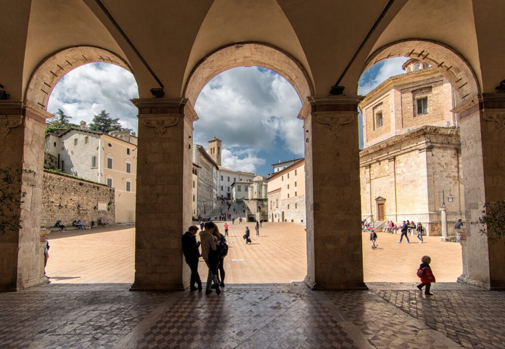 Vista della piazza antistante lil Duomo di Spoleto dall'ingresso della chiesa. Gli archi dividono scenicamente la vista sulla piazza svelando l'attività di alcune persone che passeggiano.