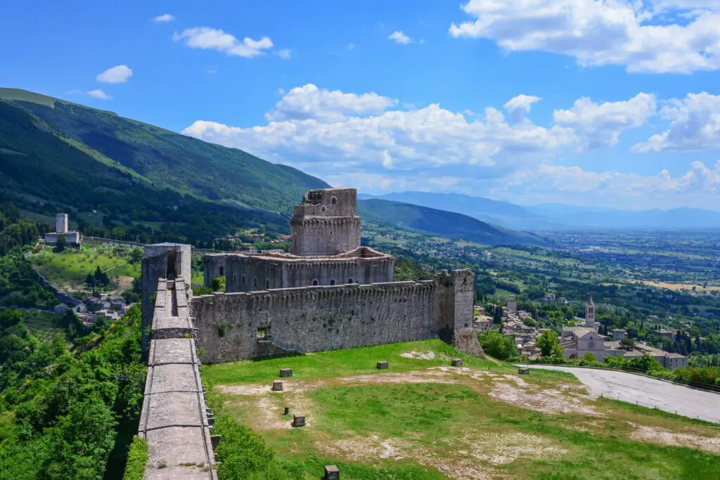 Vista delle mura della rocca maggiore. Sulla sinistra le mura che corrono dritte e poi sterzano verso destra e continuano il loro percorso. Sulla destra il verde paesaggio umbro.