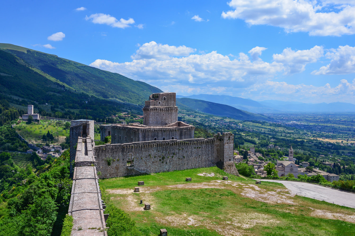 Vista delle mura della rocca maggiore. Sulla sinistra le mura che corrono dritte e poi sterzano verso destra e continuano il loro percorso. Sulla destra il verde paesaggio umbro.