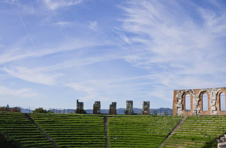 Vista della cavea e di alcune mura del Teatro Romano sovrastato da un grande cielo azzurro.