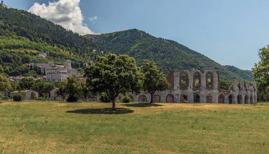 Vista del Teatro Romano nella piana con erba gialla e verde e della città di Gubbio sulle pendici del monte sullo sfondo.