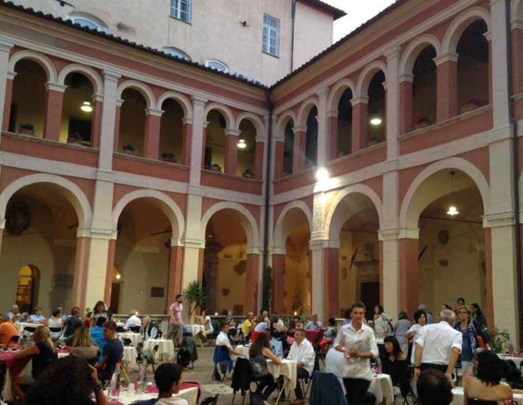 Immagine di un evento fatto nel chiostro della cattedrale di San Lorenzo. Persone sedute a dei tavoli imbanditi e persone in piedi con calici in mano con sullo sfondo gli archi del chiostro.