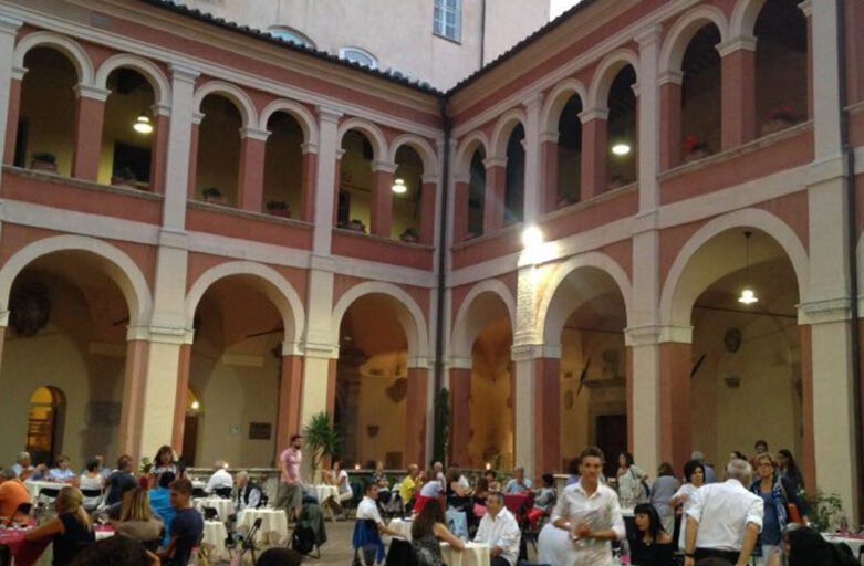 Immagine di un evento fatto nel chiostro della cattedrale di San Lorenzo. Persone sedute a dei tavoli imbanditi e persone in piedi con calici in mano con sullo sfondo gli archi del chiostro.