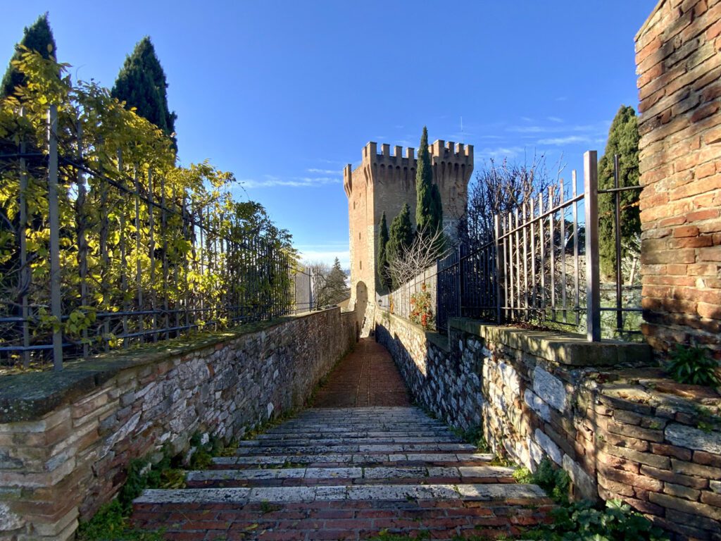 Vista laterale del cassero di Porta Sant'Angelo. In primo piano la stretta via, delimitata ai lati da bassi muretti e inferriate, che unisce l'ingresso della Torre del cassero con lo spazio antistante al tempietto, cioè la chiesa di San Michele Arcangelo. La vista della via porta al cassero, che si staglia con i suoi merli nel cielo azzurro.