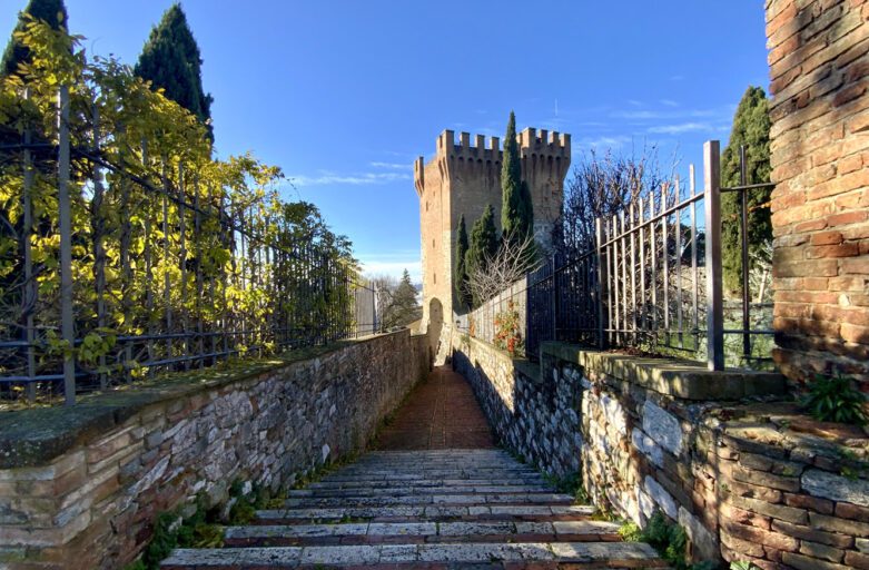 Vista laterale del cassero di Porta Sant'Angelo. In primo piano la stretta via, delimitata ai lati da bassi muretti e inferriate, che unisce l'ingresso della Torre del cassero con lo spazio antistante al tempietto, cioè la chiesa di San Michele Arcangelo. La vista della via porta al cassero, che si staglia con i suoi merli nel cielo azzurro.