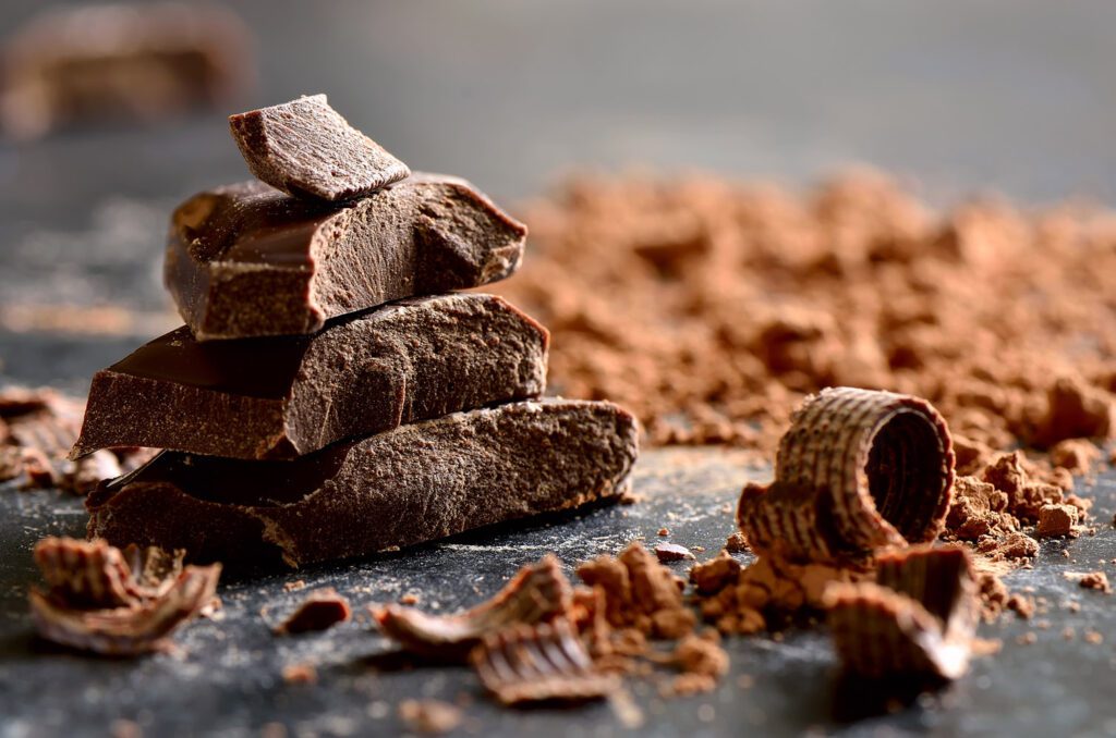 Su una superficie di colore nero sono posti polvere di cacao. riccioli di cioccolato ed una piccola pila composta da tocchi di cioccolato.