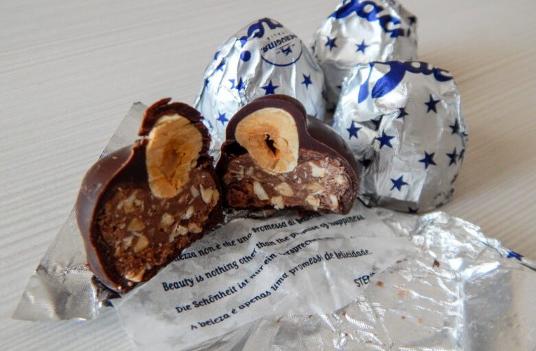 Vista di Baci Perugina. In primo piano un bacio perugino aperto a metà all'interno del quale si vede la nocciola intera e il ripieno di cioccolata. Sotto le due metà del cioccolatino è steso l'inolucro, formato da un biglietto con citazioni celebri su amore e amicizia e il classico rivestimento in alluminio argentato con stelle blu. Sullo sfondo altri due baci perugina, incartati.