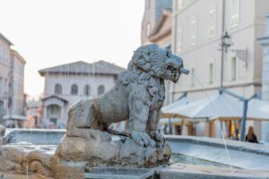 Dettaglio della fontana dei tre leoni. Vista di uno dei leoni, di profilo, dalla cui bocca esce un piccolo flusso d'acqua. Sullo sfondo la piazza.