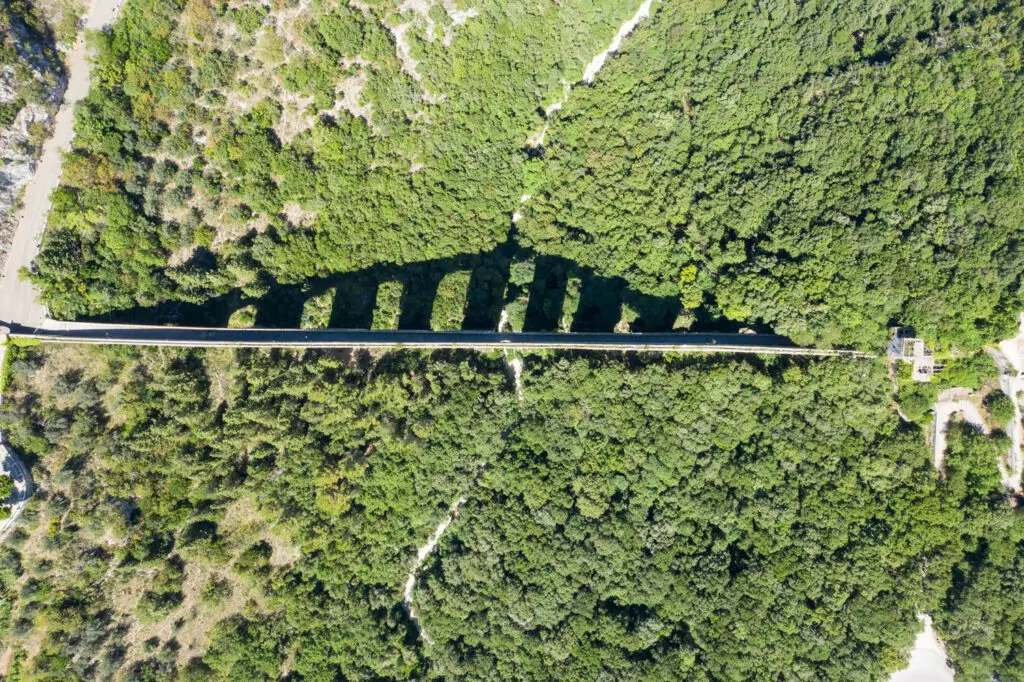 Vista aerea in verticale sul ponte delle torri. In tutta la sua lunghezza si estende tra i due monti ricoperti di boschi. Il sole proietta l’ombra degli arconi sulla vallata.