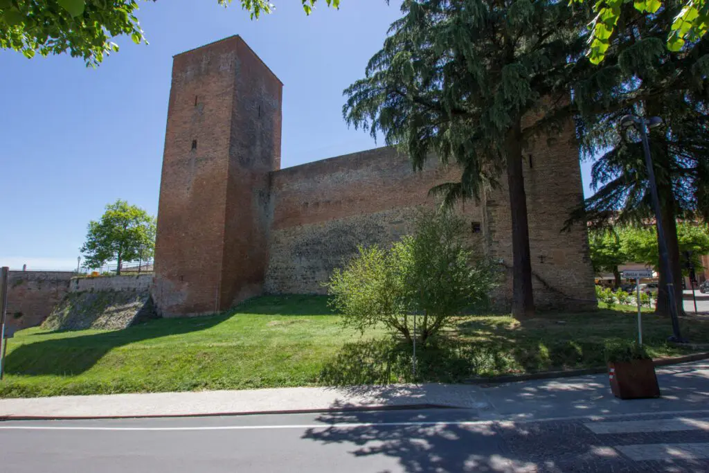 Vista del fianco della Rocca Perugina dall'esterno. Su un verde pratico si erge la rocca, del colore rosso caratteristico di città della Pieve, e si staglia nell'azzurro del cielo.