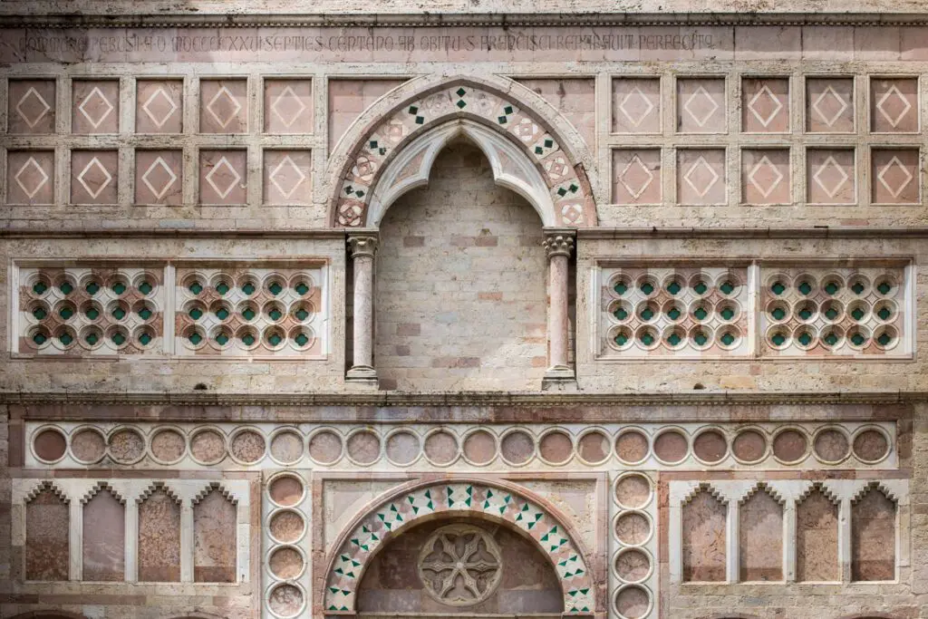 Dettaglio delle decorazioni scultore geometriche che sovrastano il portale di ingresso di San Francesco al Prato. Al centro un arco, ai cui lati, nella parte alta , si trovano cornici quadrate con rombi iscritti. Ai lati della parte bassa dell'arco decorazioni con inserzioni in pietra turchese. Sotto l'arco decorazioni a tondi che vanno a delineare i limiti del sottostante portone.