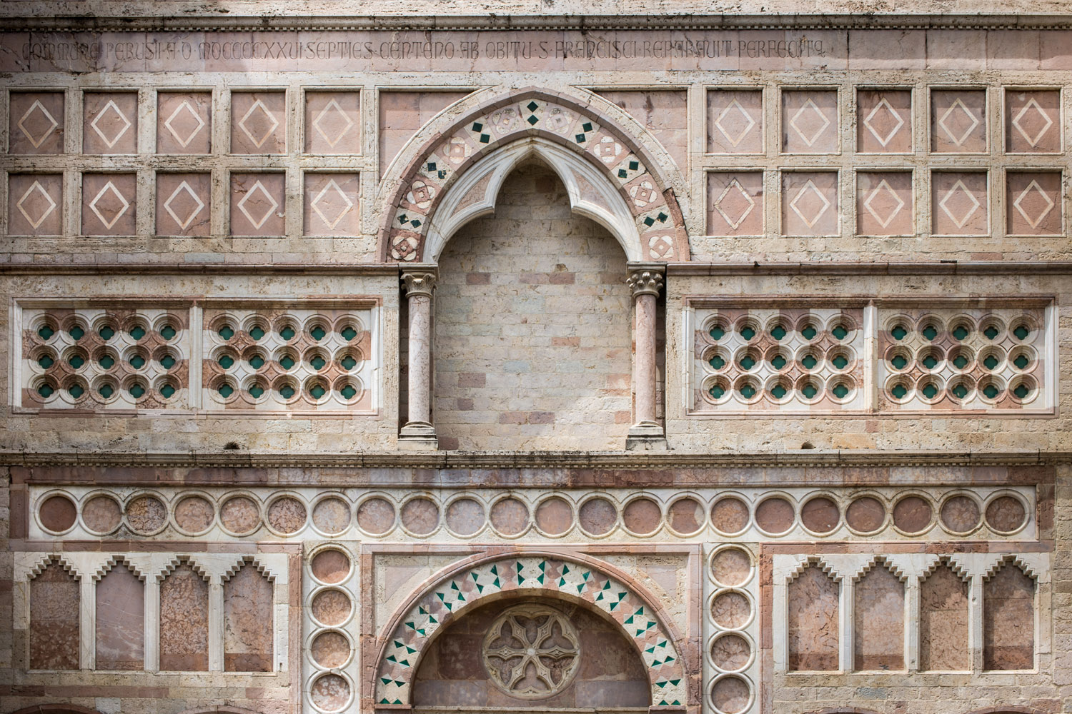 Dettaglio delle decorazioni scultore geometriche che sovrastano il portale di ingresso di San Francesco al Prato. Al centro un arco, ai cui lati, nella parte alta , si trovano cornici quadrate con rombi iscritti. Ai lati della parte bassa dell'arco decorazioni con inserzioni in pietra turchese. Sotto l'arco decorazioni a tondi che vanno a delineare i limiti del sottostante portone.