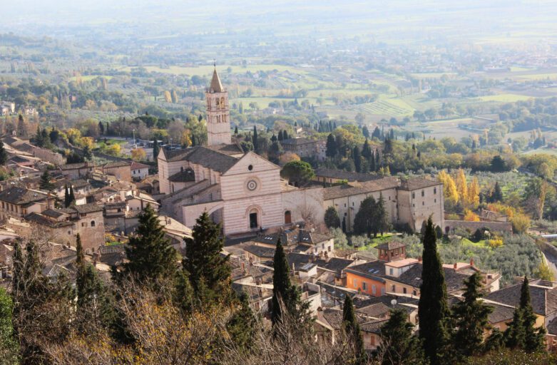 Vista panoramica della Basilica di Santa Chiara ad Assisi. Dall'abitato spuntano l'imponente facciata della chiesa e il suo campanile. Sullo sfondo le campagne umbre.