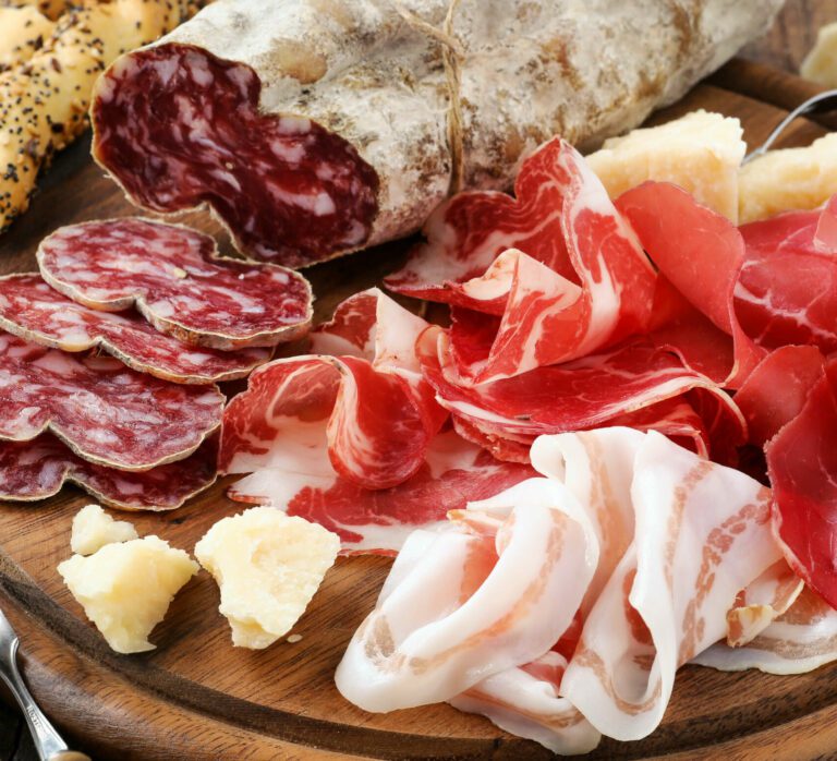 Tagliere di legno con sopra salumi affettati quali: lardo, capocollo, prosciutto e salame. Qualche pezzo di parmigiano e dei grissini sullo sfondo.