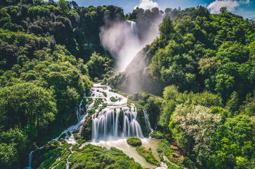 Vista frontale aerea della Cascata delle Marmore. Dal verde intenso della vegetazione salgono nuvole di vapore acqueo mentre l'acqua in tutta la sua potenza scorre verso il basso, creando diramazioni e salti.