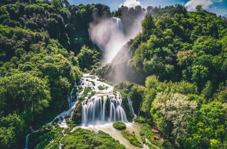 Vista frontale aerea della Cascata delle Marmore. Dal verde intenso della vegetazione salgono nuvole di vapore acqueo mentre l'acqua in tutta la sua potenza scorre verso il basso, creando diramazioni e salti.