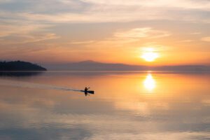 Un kayak solca le placide acque del Lago Trasimeno al tramonto. La piccola e scura figura taglia le acque diagonalmente lasciando dietro di se una scia, mentre il sole ormai arancione si avvia dietro i monti in lontananza specchiandosi nell'acqua.