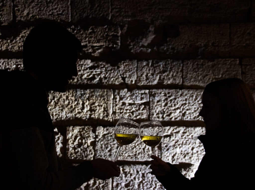 Foto frontale del muro di terrazzamento etrusco illuminato ma sfocato. In primo piano due figure scure, un ragazzo e una ragazza, si stagliano sullo sfondo del muro, mentre brindano con dei calici di vino bianco a fuoco, che risaltano staccandosi dallo sfondo.
