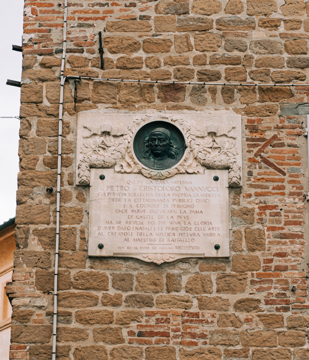 Dettaglio di un edificio antico con mattoni a vista. Apposta al muro c’è una targa di marmo con un’iscrizione in latino, che spiega che quella è la casa natale dell’artista Pietro Vannucci detto Perugino. Al di sopra della scritta, un bassorilievo con al centro il ritratto del pittore scolpito in un tondo.