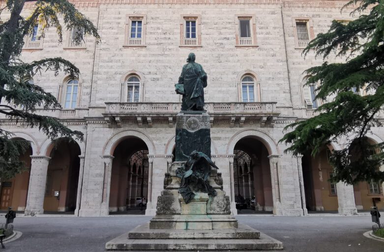 Monumento commemorativo di Pietro Vannucci detto il Perugino. La figura intera del pittore sovrasta il piedistallo impreziosito da bassorilievi; dal basso, un angelo accovacciato con in mano una tavolozza volge lo sguardo verso l’artista. La statua è in mezzo a un giardino con delle panchine piazzate sotto dei grandi abeti. Sullo sfondo, un grande edificio storico con una balconata nella parte centrale della facciata.