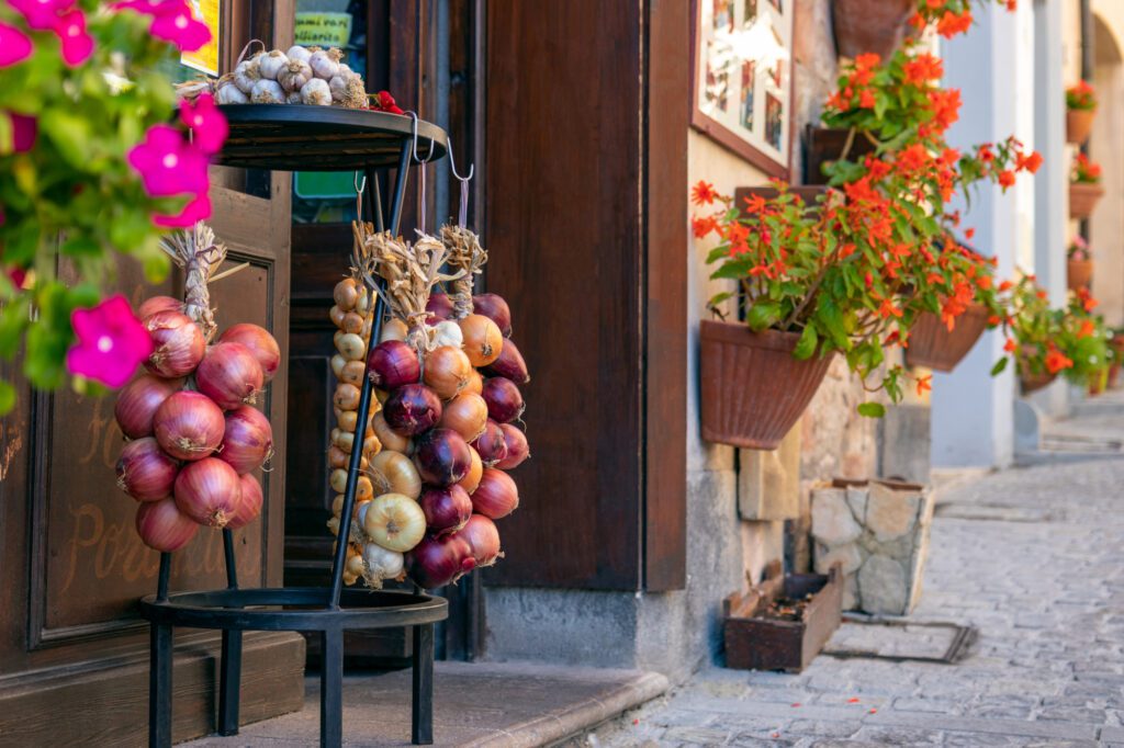 veduta esterna parziale di un negozio che espone fuori trecce di cipolle di tre varietà diverse: borettana, rossa e dorata. Il negozio si affaccia su un vicolo di Cannara addobbato con vasi di fiori colorati.