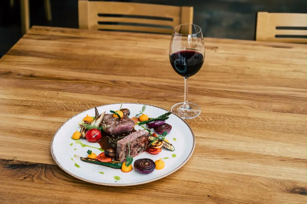 vista frontale dall’alto di una tavola da pranzo con un piatto di carne rossa condita con verdure varie. Accanto al piatto c’è un calice di vino rosso Sagrantino.