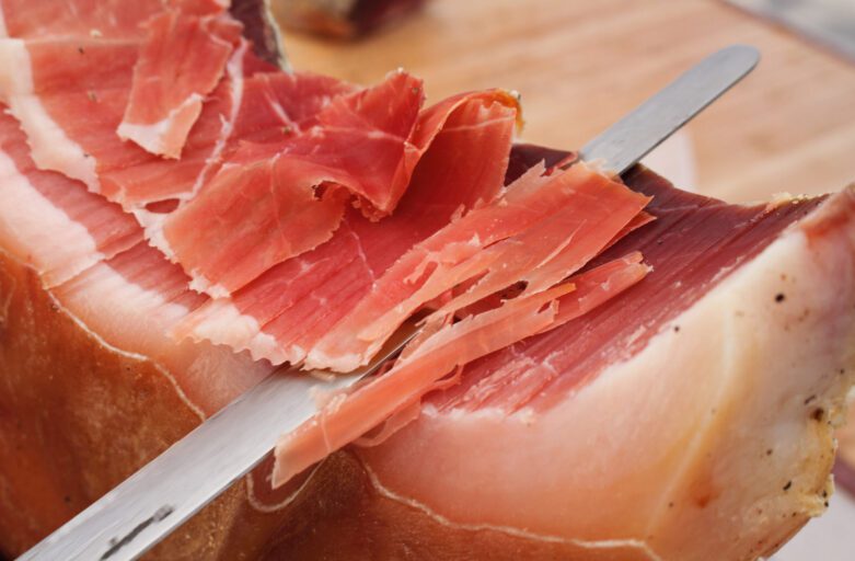 immagine ravvicinata di una fetta di prosciutto di norcia che viene affettata a mano con un coltello.