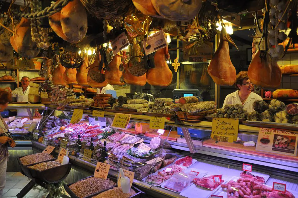 immagine dell’interno di una salumeria a Norcia. Nella parte in basso e centrale di tutta l’immagine si trova un bancone refrigerante, tipico delle salumerie, dove sono esposte alcune specialità in vendita, per esempio formaggi, salumi di vario genere (salame, capocollo, carne cruda). Sopra il bancone sono esposte diverse tipologie di salami (per esempio salame al cinghiale, salame di Norcia ecc.). Nella parte superiore dell’immagine sono raffigurati i prosciutti di Norcia appesi al soffitto del negozio.