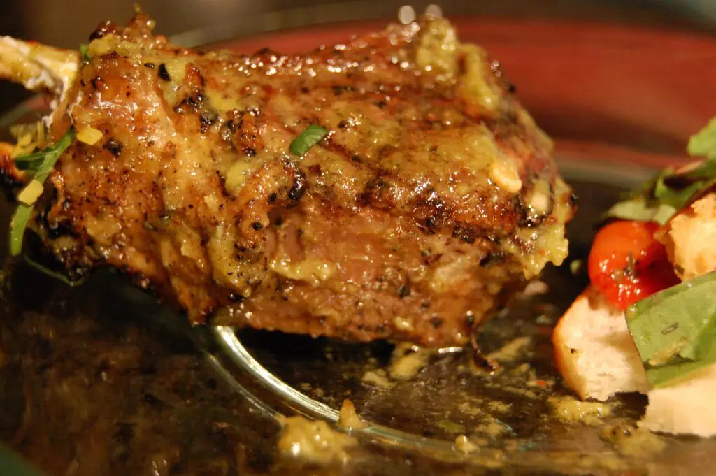 inquadratura ravvicinata su delle costolette di agnello cotte alla griglia sul lato sinistro dell’immagine. Sul lato destro si intravedono, in modo sfocato, delle verdure a condimento, come insalata e pomodori, accompagnati da del pane.