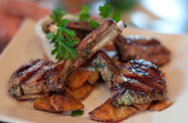 inquadratura ravvicinata su un piatto con delle costolette di agnello adagiate su patate cotte al forno, il tutto condito con del prezzemolo.