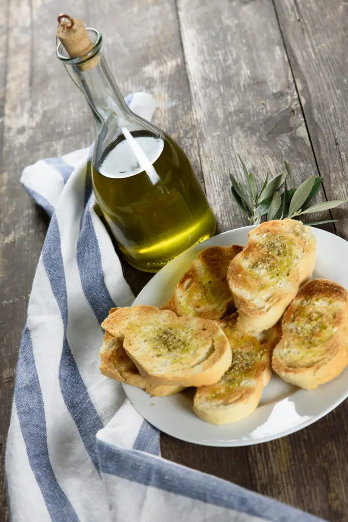 inquadratura laterale dall’alto di una tavola dove sono adagiati: sulla sinistra, un canovaccio da cucina a righe blu e bianche. Accanto, più verso il centro del tavolo, un piatto con alcune fette di bruschette condite con olio. Tra il canovaccio e il piatto, è appoggiata una bottiglia di vetro che contiene dell’olio extravergine di oliva. 