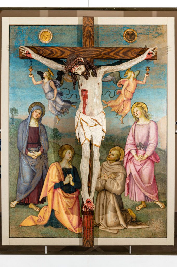 visione d’insieme della faccia anteriore della Pala di Monteripido. È una fusione tra pittura e scultura perché su questa faccia è stata collocata una statua in legno di Cristo crocifisso. La scena del Calvario è completata da quattro figure dipinte dal Perugino ai piedi della Croce. In piedi, ci sono la Vergine (a sinistra) e San Giovanni Evangelista (a destra). Inginocchiati, invece, ci sono Maria Maddalena (a sinistra) e San Francesco d’Assisi (a destra). Nella parte superiore della scena, due angeli sono in volo ai lati di Cristo ed entrambi con un calice raccolgono il sangue che cade dalle sue mani ferite. Sopra l’asse trasversale della croce, sono dipinti in cielo il Sole e la Luna.