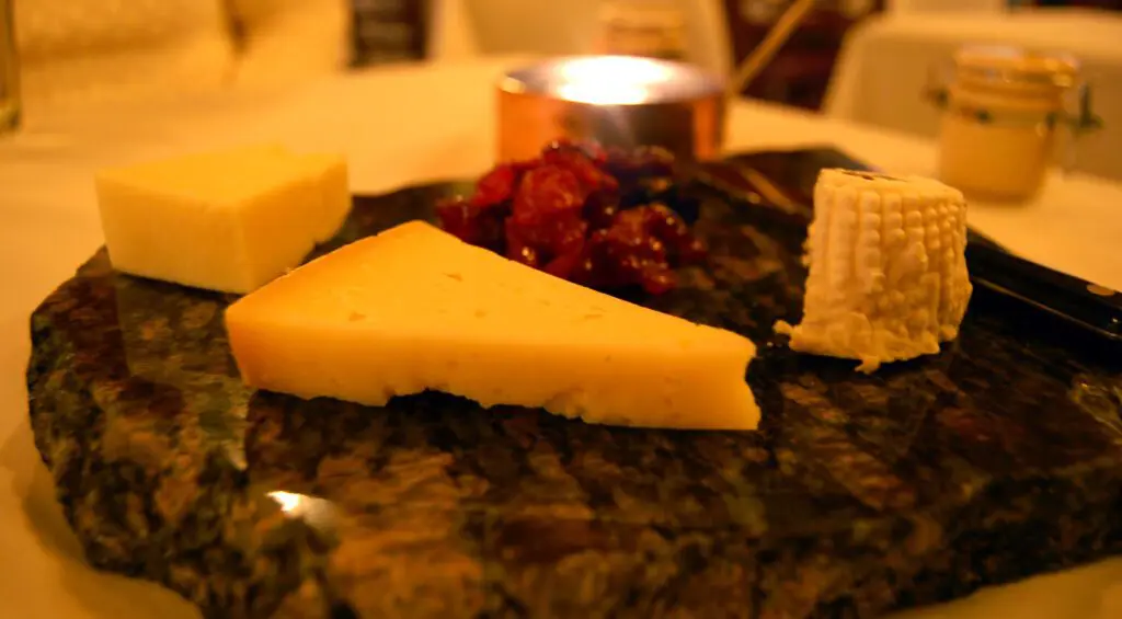 inquadratura ravvicinata e laterale di un tagliere da degustazione in marmo dove sono adagiati spicchi di vari tipi di formaggi. In primo piano si trova una fetta di pecorino toscano; ai lati della fetta ci sono altri due tipi di formaggi e al centro del tagliere delle verdure di accompagnamento.