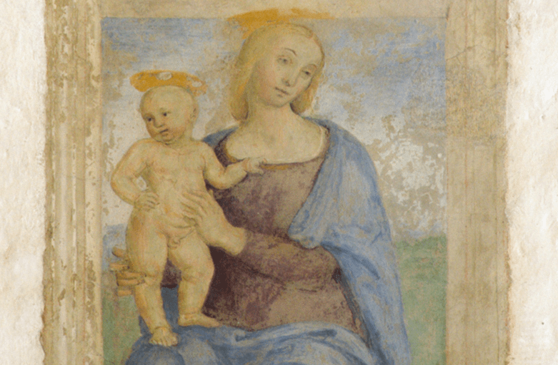 Dettaglio dell'affresco raffigurante una Madonna seduta su un banco di legno, che sembra sporgere dal muro, ritratta nell’atto di sorreggere il Bambino in piedi sul suo ginocchio destro. Il Bambino è aggrappato con la mano sinistra alla tunica della madre, come per tenersi meglio in piedi. Nella parte bassa dell’affresco compare l’iscrizione “Agniolus Toni Angeli fecit fieri MDXXII”.