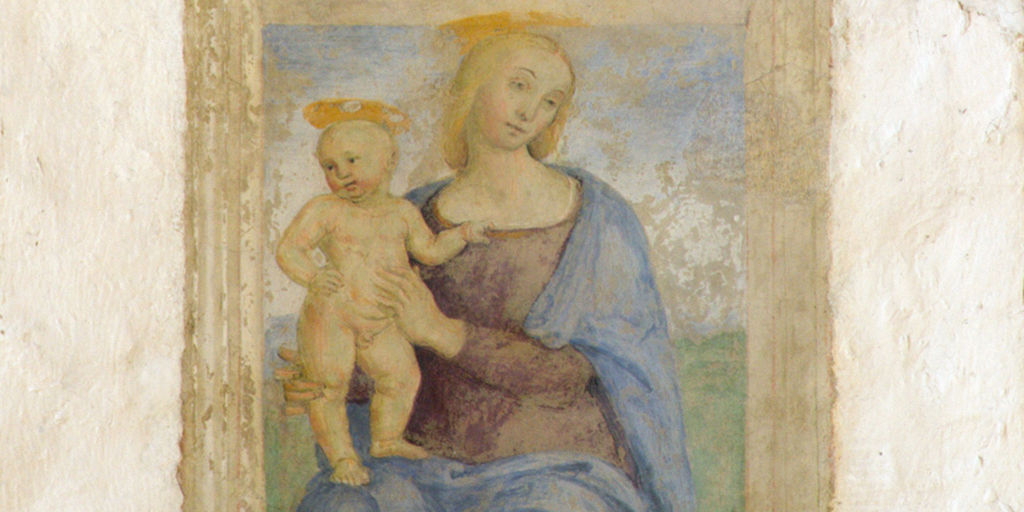 Dettaglio dell'affresco raffigurante una Madonna seduta su un banco di legno, che sembra sporgere dal muro, ritratta nell’atto di sorreggere il Bambino in piedi sul suo ginocchio destro. Il Bambino è aggrappato con la mano sinistra alla tunica della madre, come per tenersi meglio in piedi. Nella parte bassa dell’affresco compare l’iscrizione “Agniolus Toni Angeli fecit fieri MDXXII”.