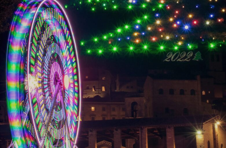 vista frontale e dal basso dell’albero di Natale di Gubbio. La sagoma di luci a led che compongono la figura dell’Albero di Natale si trova sulle pendici del Monte Ingino, sopra la città di Gubbio. Accanto alla punta dell’albero si intravede la parte superiore della Basilica di Sant’Ubaldo. La foto è stata scattata di notte e le luminarie sono accese. Nella parte centrale dell’immagine ci sono gli scorci della città sotto la base dell’albero di luci. Sulla sinistra in primo piano, c’è una ruota panoramica illuminata.