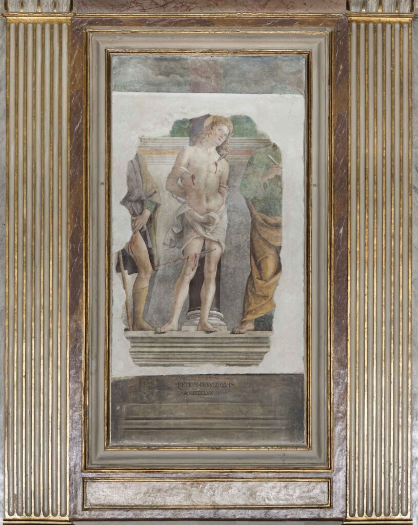 Overall view of the fresco. In the centre, there is Saint Sebastian tied to a column, his head and gaze turned upwards and his body is pierced by three arrows, two on his chest and one on his right leg. He is completely naked, except from a cloth covering his hips. On the left, there is part of a human figure showing a bleeding wound on the left inner thigh (presumably St. Roch) wearing a short tunic and another one on the right wearing a long tunic holding a key in his right hand (probably St. Peter).