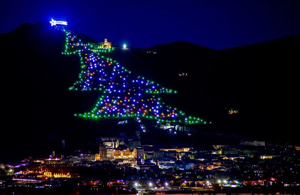 vista panoramica laterale dell’Albero di Natale di Gubbio del 2019. L’albero illuminato si trova sulle pendici del Monte Ingino. Sulla sommità dell’Albero si trova la Stella Cometa; accanto è illuminata la Basilica di Sant’Ubaldo. Alla base dell’Albero si vedere una panoramica dall’alto della città di Gubbio.