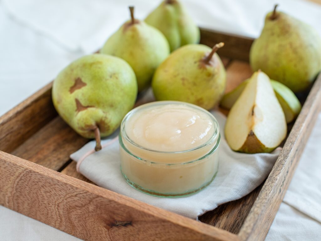 in primo piano nell’immagine c’è un piccolo vasetto di vetro che contiene marmellata di pera di monteleone di orvieto. Sullo sfondo sfocate sono adagiate delle pere, sia intere che tagliate a metà. Il tutto è appoggiato su un vassoio di legno con i bordi rialzati.