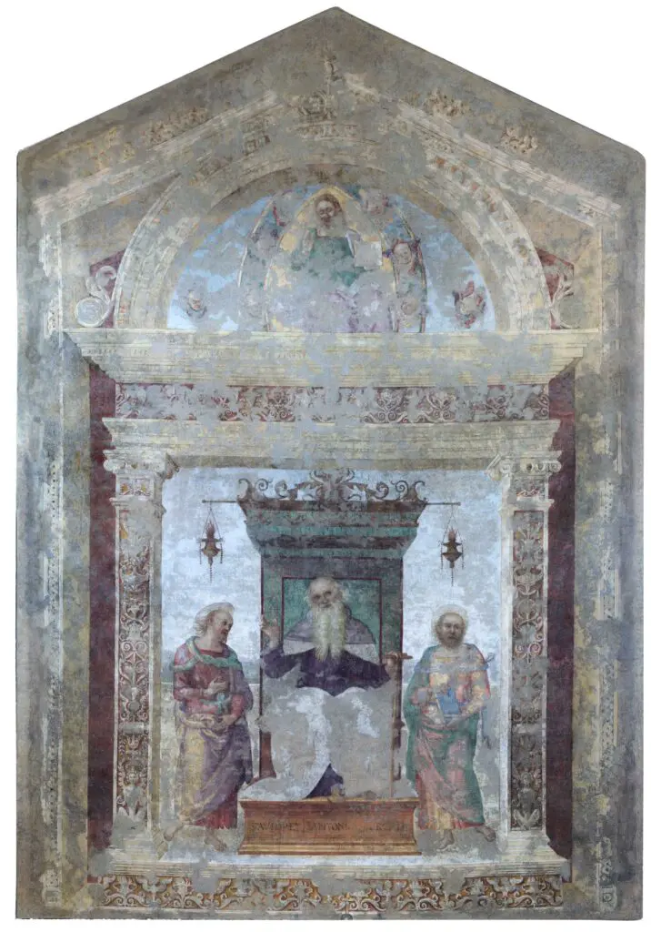 Immagine dell’affresco del Perugino intitolato “Sant'Antonio Abate tra i Santi Paolo Eremita e Marcello” e conservato all’interno della chiesa dei Santi Pietro e Paolo a Città della Pieve. Nella scena principale, Sant’Antonio Abate è dipinto al centro, seduto su un elegante trono rialzato da cui pendono due lanterne. Mentre volge uno sguardo intento all’osservatore, il Santo ha la mano destra sollevata e la mano sinistra appoggiata al bastone, un oggetto con cui viene solitamente raffigurato nelle pitture sacre. Sant’Antonio Abate è affiancato da San Paolo Eremita e San Marcello, come indica la scritta alla base del trono. Nella lunetta che sormonta la scena principale è raffigurato Cristo benedicente circondato dai putti, bambini nudi alati presenti nella maggior parte delle opere sacre del Perugino. Nel 1861, il terremoto ha colpito la Chiesa e danneggiato l’affresco. L’opera è stata quindi trasferita in una tela e restaurata, ma non è stato possibile riportarla allo stato originale date le cadute di colore in gran parte dell’opera.